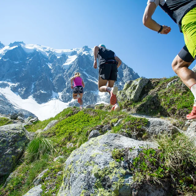 Three trail runners running up a steep trail towards the mountains in the Alps